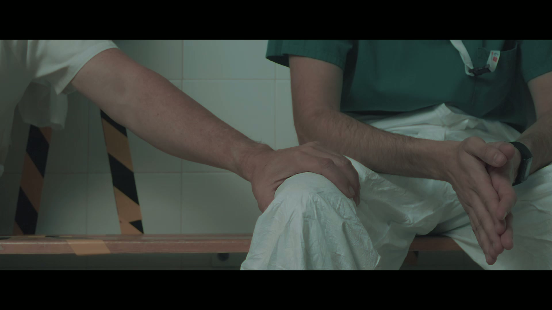 Fotograma de 5min: la mano de un compañero sobre la rodilla de un sanitario sentado, con las manos entrelazadas